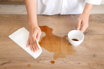 Woman wiping spilled coffee with paper towel on table in kitchen, closeup