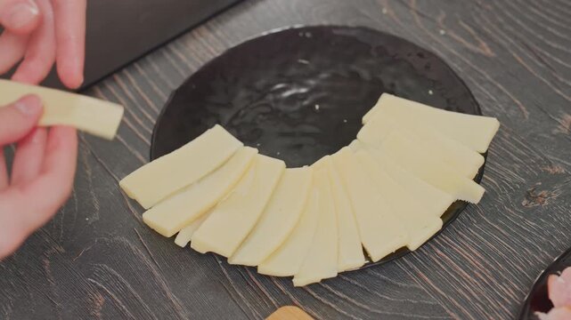 close up of chef using hands to arrange yellow cheese slices in circular pattern on black ceramic plate placed on wooden table, adjusting plate position while organizing dairy pieces with care
