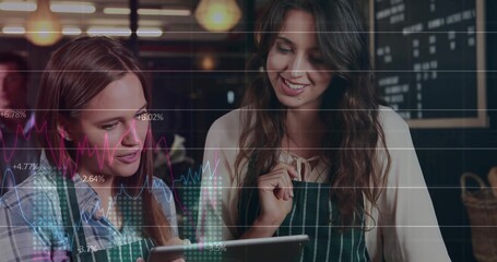 Examining tablet two baristas wearing green striped aprons at cafe counter, with chalkboard menu