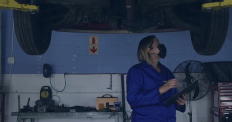 Inspecting mechanic taking notes under raised car in repair bay, with clipboard and fan, copy space