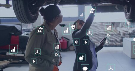 Using tablet, two female mechanics in coveralls inspecting car underbody in workshop, overlay icons