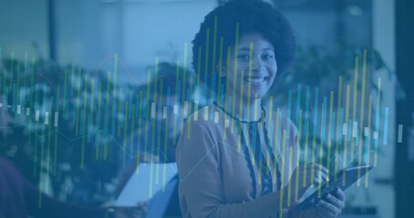 Woman in tan sweater and lanyard holding tablet and analyzing data graphs in office with laptops