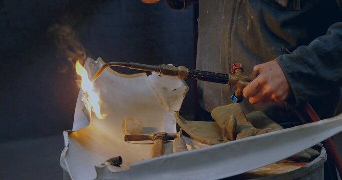 Shaping metal panel metalworker wearing jacket using blowtorch on drum in shop, with hammers mallet