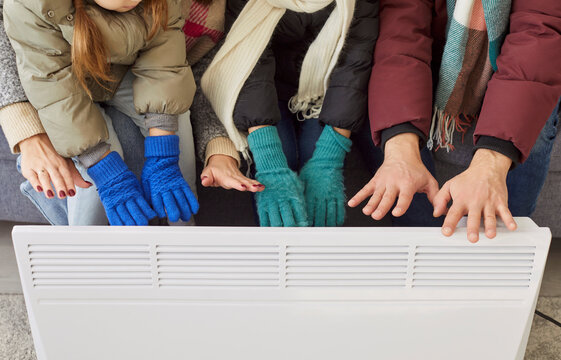 Top view of a family hands on electric heater warming up in chilly room. Frozen parents with children in winter outwear are freezing suffering from heating problems. People getting cold at home.