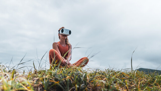 Female in sportswear practicing relaxation with VR headset on grass. Combination of modern technology, meditation, and outdoor lifestyle. Perfect for concepts of future health and virtual wellness.