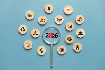 Magnifying glass focusing on 2026 target surrounded by wooden business icons representing New Year goals, strategic planning, OKR, KPI, action plan, innovation, AI, sustainability, and business growth