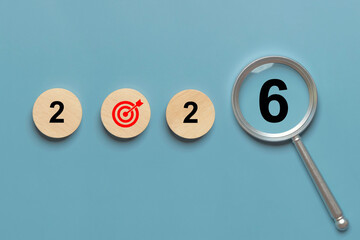 Magnifying glass focuses on the number 6 to complete 2026 with wooden discs and a red target on blue background, for New Year goals, annual planning, OKR, KPI, action plan, strategy, focus, and growth