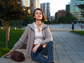 A stylish young woman sits on a park bench in an urban setting during golden hour. She wears a...