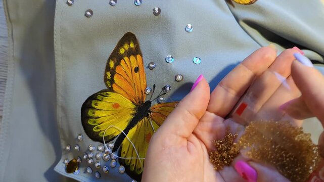 Custom hand-painted butterfly with beads and sequins on textile. Artistic fashion design and personal creativity concept. Female hand holding golden beads for decoration.
