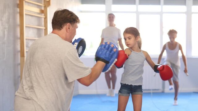 Concentrated sporty tween girl in boxing gloves practicing self-defense techniques with family in gym, throwing punches on focus mitts in hands of father