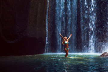 Woman stand in underground cave pool under falling fresh water of Tukad Cepung waterfall. Nature day tour, hiking activity adventure and fun at family tourist camp on summer vacation in Bali island