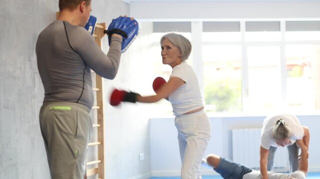 Senior lady and male couch are boxing in gym, man helps student to work out force of blow. Modern positive family chooses active hobby