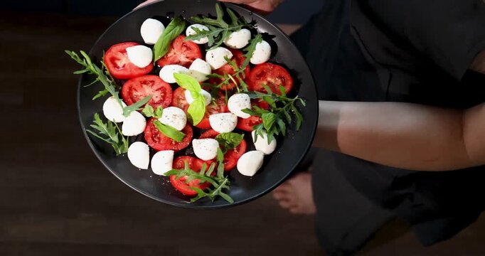 Someone is holding a black plate. The plate holds a Caprese salad with sliced tomatoes, mozzarella balls, and fresh herbs. It is a bright, sunny day.
