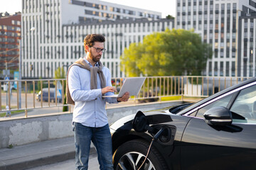 A man uses a laptop while his electric vehicle charges, showcasing modern technology and sustainable transportation in an urban setting.