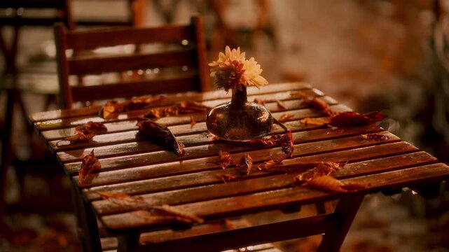Raindrops gently hit a wooden table surrounded by colorful autumn leaves. A simple flower vase adds charm to the cozy outdoor setting on a rainy day.
