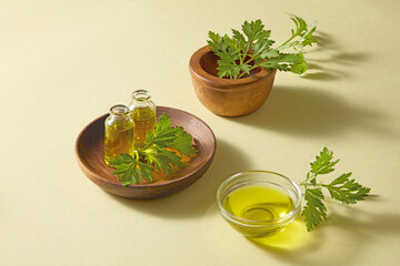 Wooden bowl filled with mugwort leaves and glass of oil, styled for skincare cosmetic raw ingredient photography presentation.