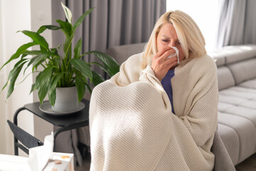 Blonde woman covered in cozy plaid sitting in living room, blowing her nose with tissue. Concept of cold, flu, or covid infection, home healing and relaxation.