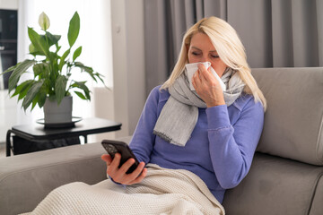 Middle aged blonde woman feeling unwell, sitting on couch and using her smartphone for work.
