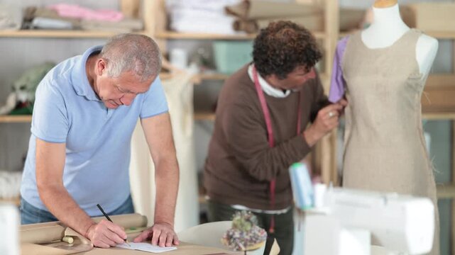 Mature tailor draws a sketch and pattern of clothes on a brown paper next to the assistant. Process of creating clothes in a sewing workshop. High quality 4k footage