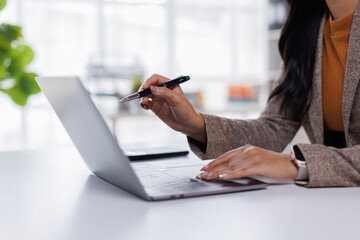 Business asian woman using laptop computer and holding a pen while working at her desk in a bright office
