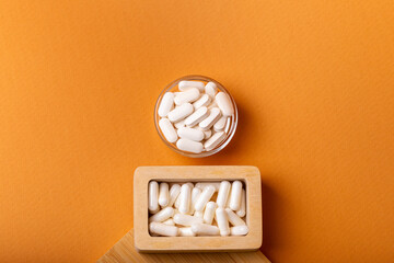 White vitamin pills in wooden bos and glass bowl from above on a wooden desk on orange bakcground.