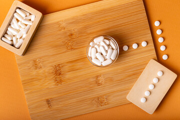White vitamin pills in wooden bos and glass bowl from above on a wooden desk on orange bakcground.