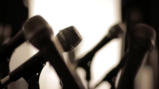 A line of professional microphones, one in focus, silhouetted against a softly lit background