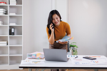 Young professional asian woman on a phone call, reviewing documents at a stylish office desk during daylight hours.