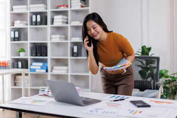 Young professional asian woman on a phone call, reviewing documents at a stylish office desk during daylight hours.