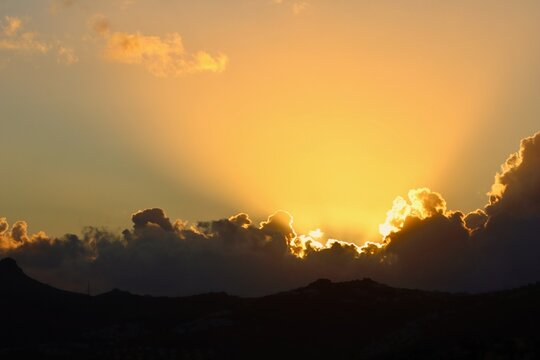 A spectacular sunset or sunrise scene with intense golden crepuscular rays bursting from behind dark, heavy cumulus clouds, silhouetting the hilltops and filling the sky with warm colors.