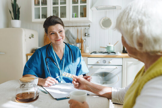Smiling caring healthcare worker in blue uniform answering questions of her aged female patient, retired woman showing her blister with pills while sitting in kitchen. Consulting with doctor at home