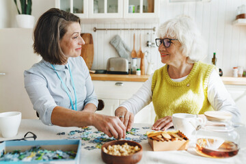 Senior lady in glasses solving puzzle with assistance of her caregiver sitting at kitchen table,...