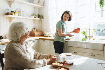 Caring mature female caregiver helping her senior patient to cook, standing next to counter holding...