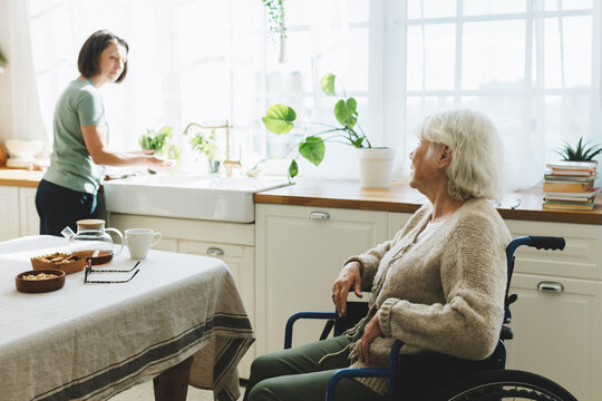Senior lady in wheelchair in kitchen talking to nurse cooking dinner or washing dishes. Social worker helping retired disabled female with domestic chores at home, having warm conversation