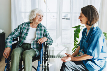 Obraz premium Two people in retirement house having conversation against panoramic window, caregiver counseling her senior patient with physical disability sitting in wheelchair and listening to her complaints