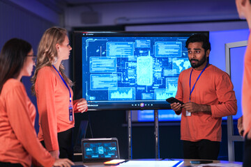 In a government command center, a diverse team of intelligence analysts collaborates on a critical mission, analyzing real-time data from a tablet, phone, and a large video wall.