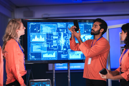 In a government command center, a diverse team of intelligence analysts collaborates on a critical mission, analyzing real-time data from a tablet, phone, and a large video wall.