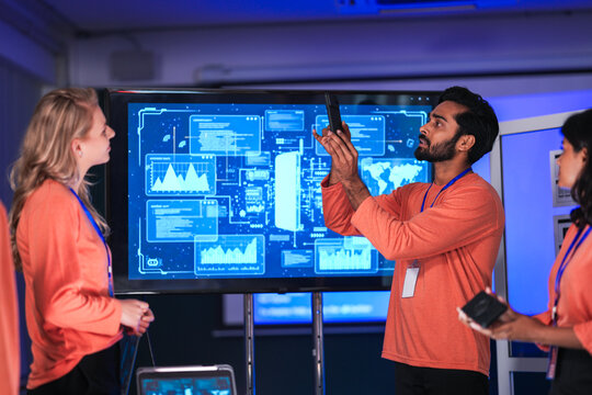 In a government command center, a diverse team of intelligence analysts collaborates on a critical mission, analyzing real-time data from a tablet, phone, and a large video wall.