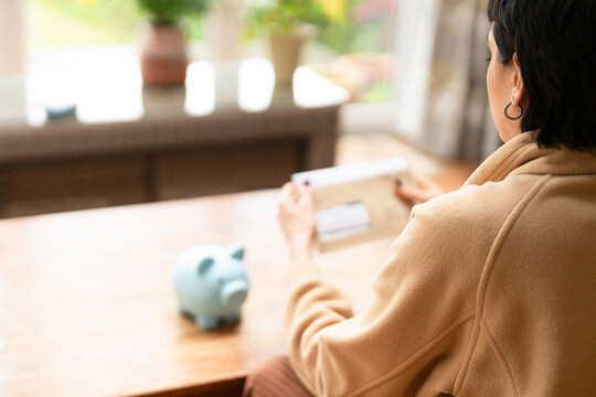 Person sitting at a wooden table, holding a document while looking at a piggy bank in a cozy indoor setting - Powered by Adobe