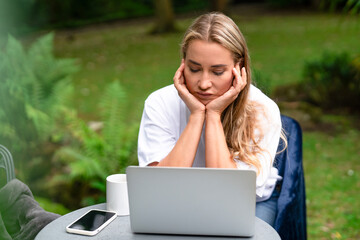Young woman working on laptop outdoors, feeling stressed while surrounded by greenery during a calm...