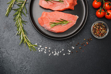 Raw veal steaks on black ceramic plate with herbs tomatoes and spices on kitchen tabl