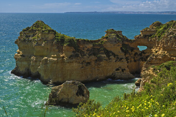Fototapeta premium coastal view with golden cliffs, sandy beach, and turquoise ocean waves. The cliffs feature layered rock and sparse vegetation, with buildings visible on top in Portimao, Algarve, Portugal