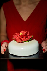 The woman presents an elegantly crafted cake featuring a vibrant flower on top. The surroundings are softly lit, enhancing the festive atmosphere of the occasion.