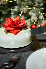 A person is cutting a festive cake adorned with a red flower, surrounded by christmas holiday decorations, glowing lights, and a joyful atmosphere perfect for winter celebrations.