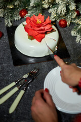 A person is cutting a festive cake adorned with a red flower, surrounded by christmas holiday decorations, glowing lights, and a joyful atmosphere perfect for winter celebrations.