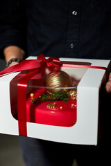 Someone holds a red gift box wrapped in white with a red ribbon, featuring a golden ornament and festive decorations, perfect for the holiday season.