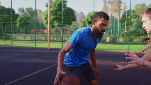 Portrait of skillful active sporty fit African American male basketball player receiving pass from teammate, making three points shot while multi ethnic athletes playing streetball on outdoor court.