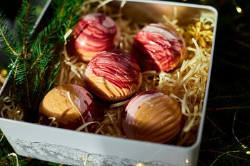 An elegant assortment of colorful dessert spheres is beautifully arranged in a festive gift box filled with straw and surrounded by fresh greenery and decorative touches, perfect for celebrations.