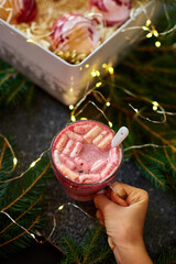 A delicious holiday dessert drink features a rich pink color, topped with fluffy marshmallows, as it is held against a backdrop of festive decorations and soft lights.