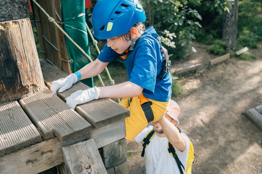 Boy climbing onto wooden platform with parent support in forest park
